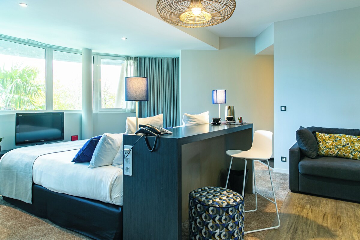 Ein modernes Hotelzimmer mit einem Bett, einem Schreibtisch, einem Sessel, einer Couch und einem Fernseher.