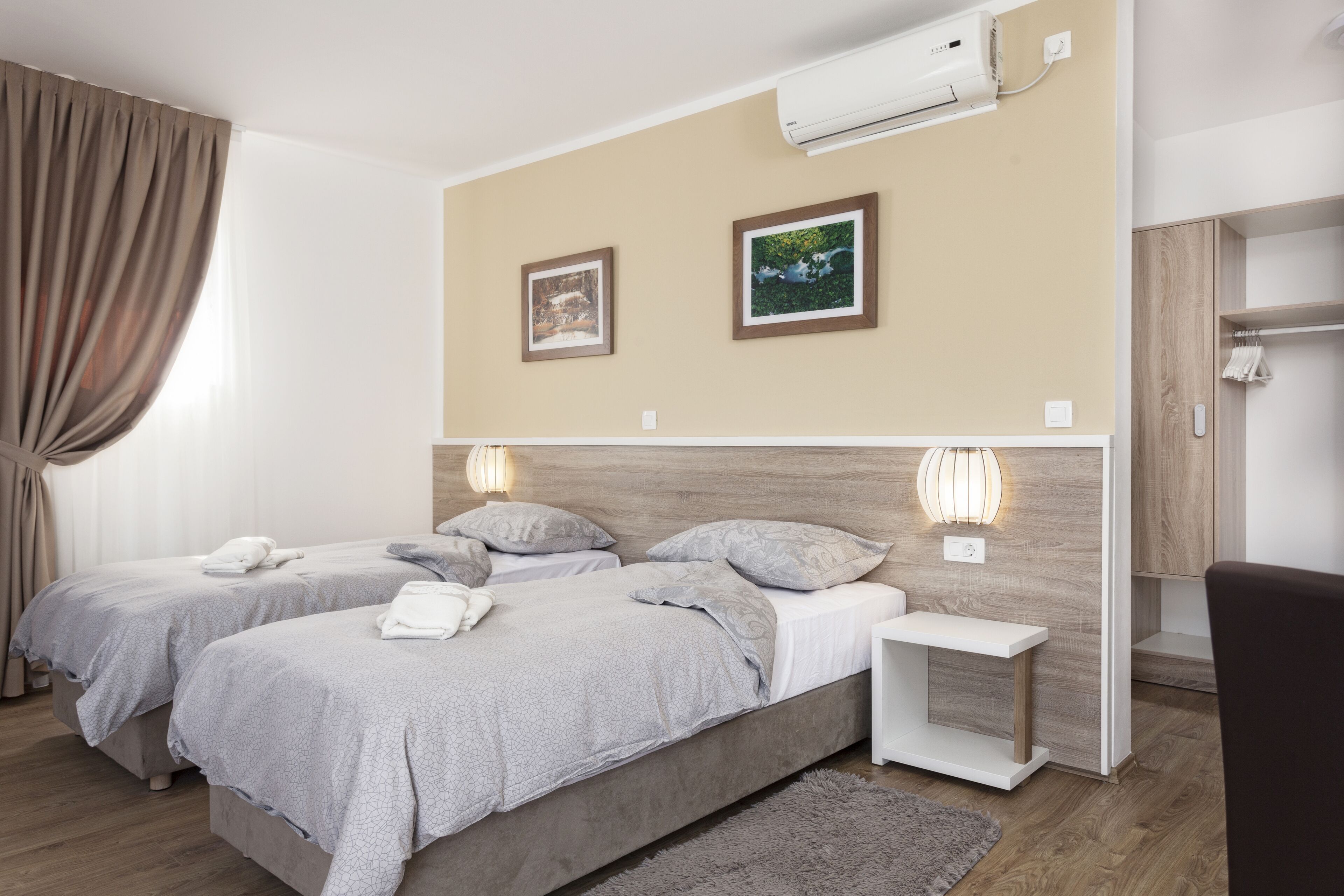 A hotel room with two beds, a wooden headboard, a small bedside table, a wall-mounted air conditioner, and two framed pictures.