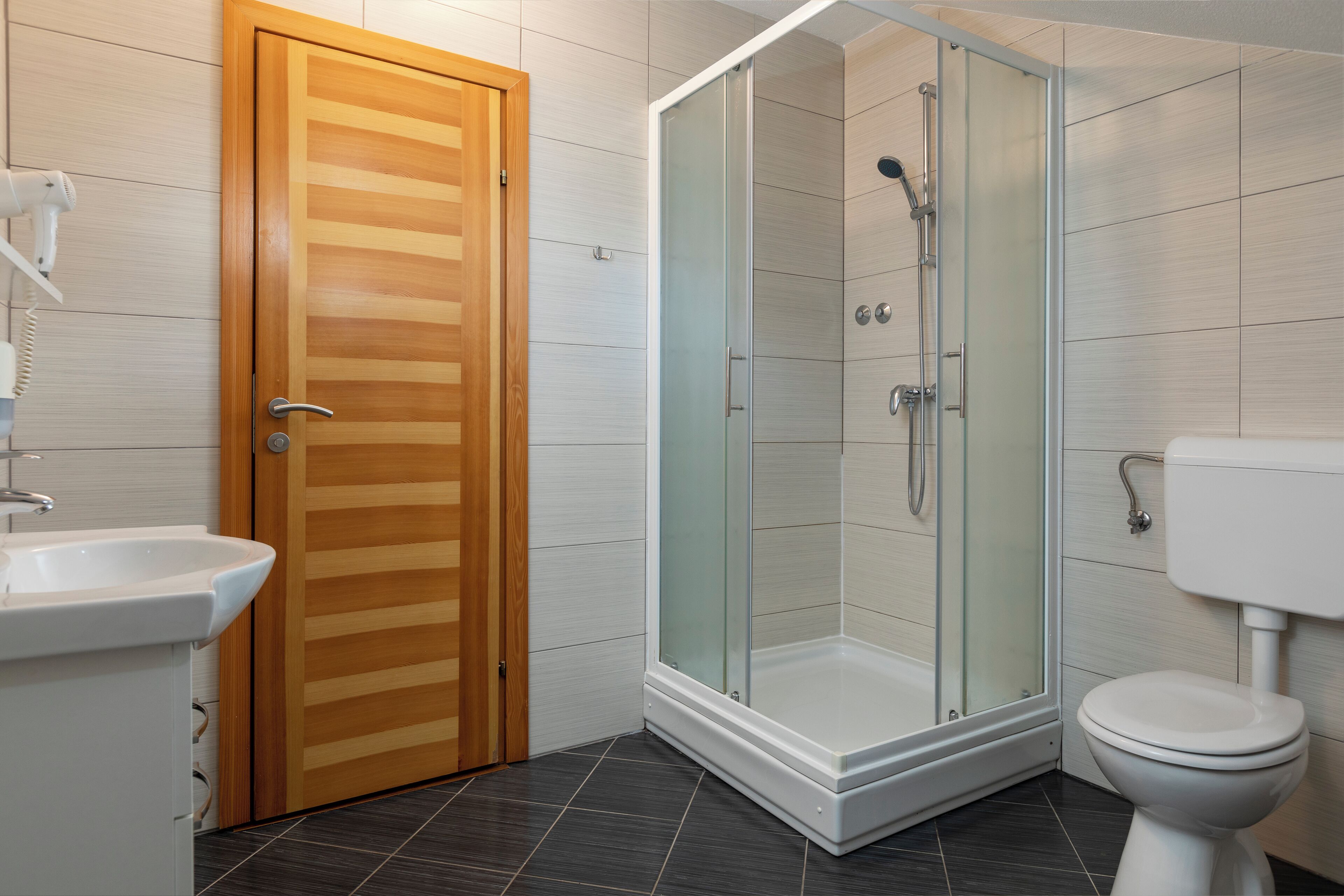 Un baño moderno con puerta de madera, cabina de ducha con paredes de vidrio, lavamanos blanco y un inodoro.