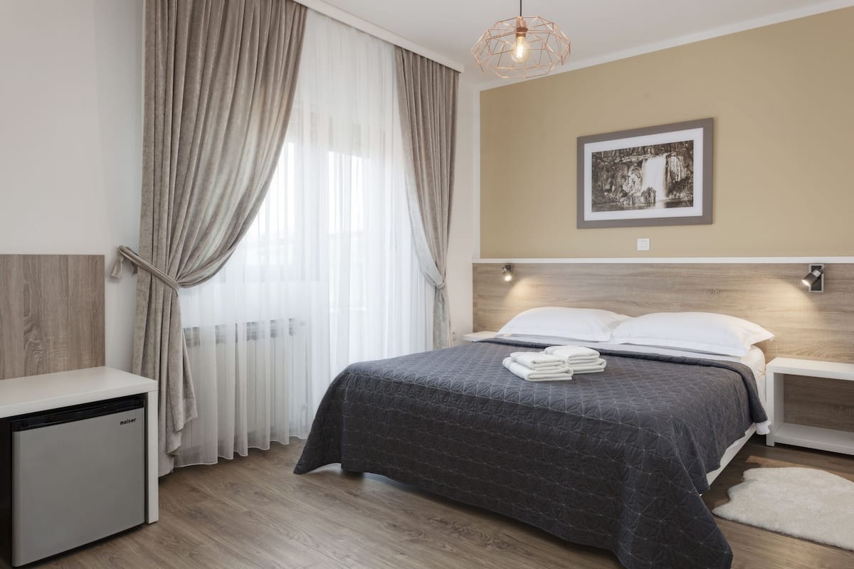 A modern bedroom with a bed, bedside tables, a small refrigerator, and a window with curtains.