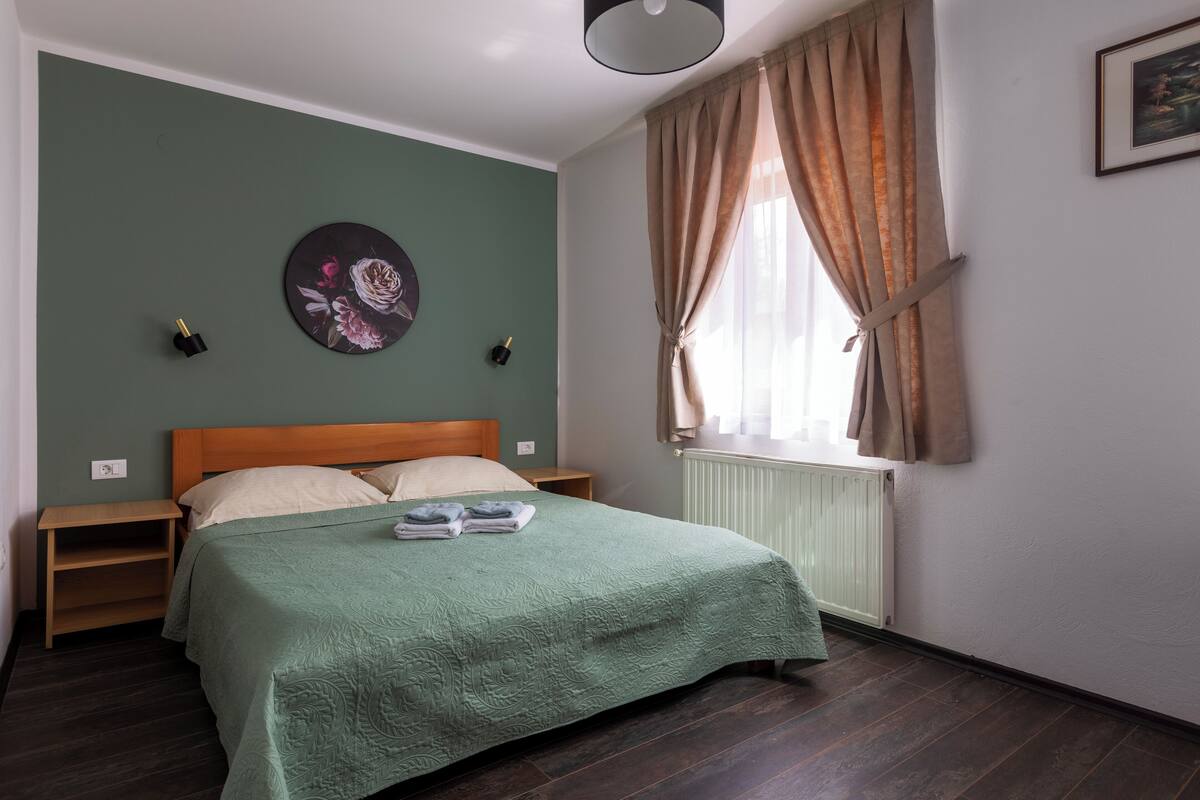 A bedroom with a green bedspread, a wooden headboard, a nightstand, a wall clock, and a window with curtains.