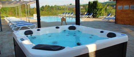 A hot tub with a pool in the background, lounge chairs, and a wooden cabin.