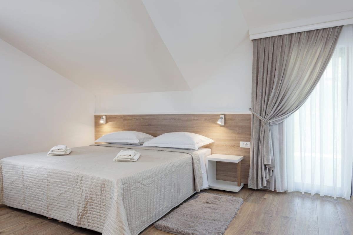 A modern hotel room with a large bed, a bedside table, a nightstand, and a window with curtains.