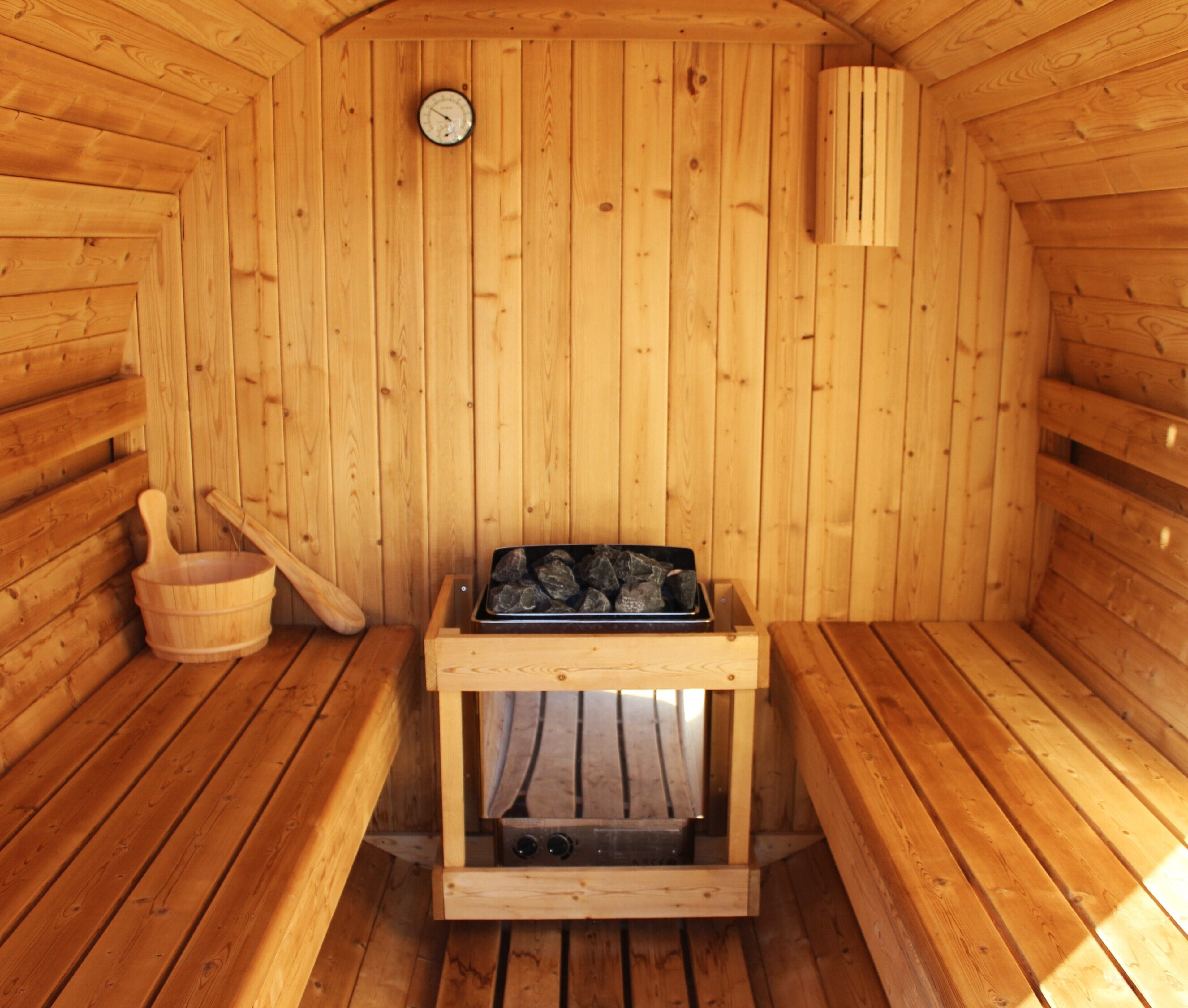 A sauna room with wooden walls, a wooden bench, a wooden bucket, a sauna heater, and a thermometer on the wall.