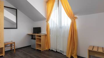 Una habitación con techo inclinado, un escritorio de madera con televisor, una silla pequeña y una ventana con cortinas amarillas.