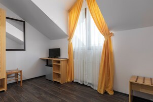 Una habitación con techo inclinado, un escritorio de madera con televisor, una silla pequeña y una ventana con cortinas amarillas.