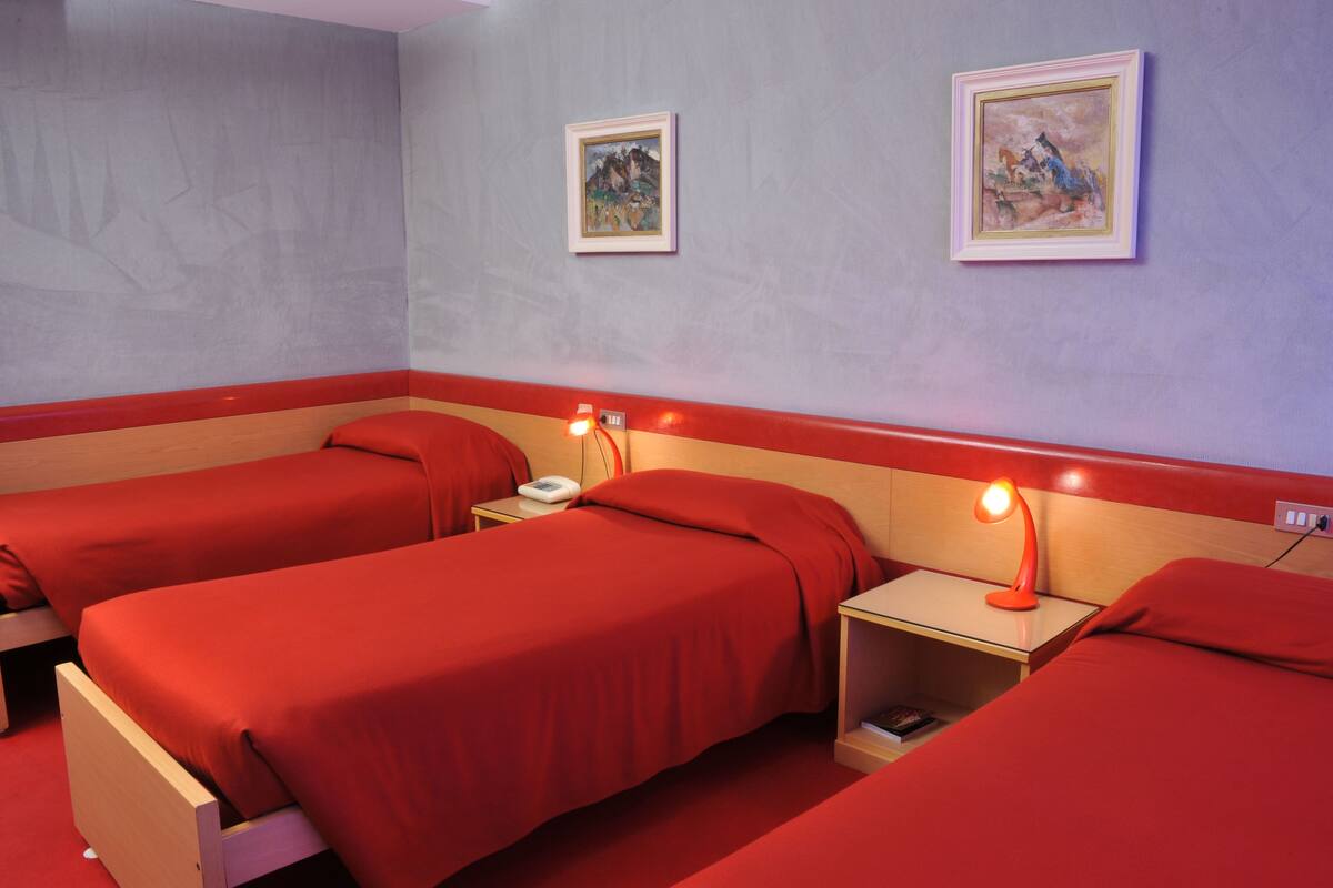 Quarto de hotel com duas camas de solteiro, cada uma com colcha vermelha e mesa de cabeceira com abajur. Há duas quadros na parede.
