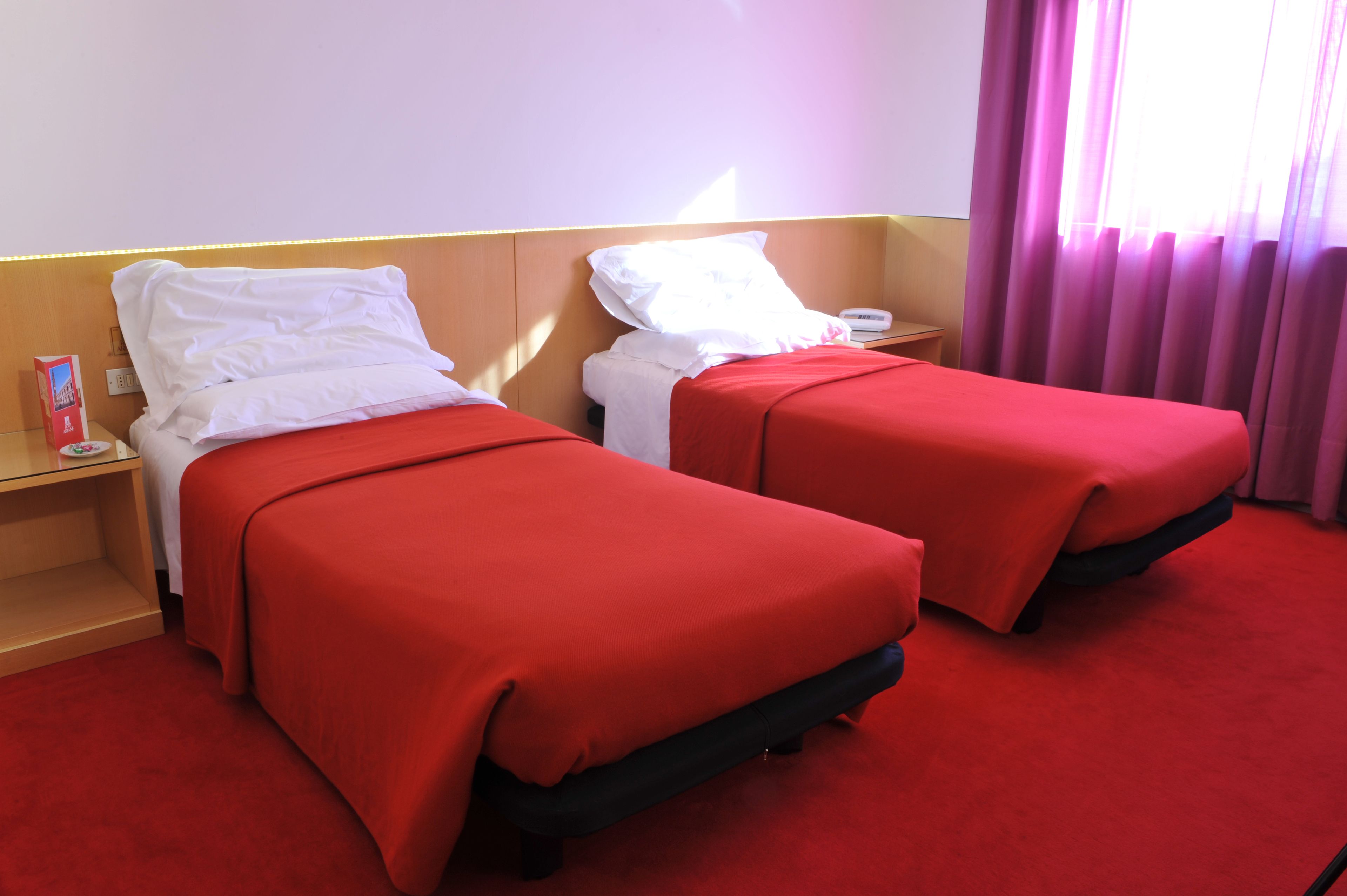 A hotel room with two beds, each with a red bedspread and white pillows. There is a wooden headboard, a nightstand with a lamp, and a window with purple curtains.