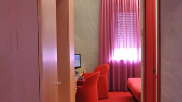 A hotel room with a red carpet, a desk with a computer, a bed, and a window with pink curtains.