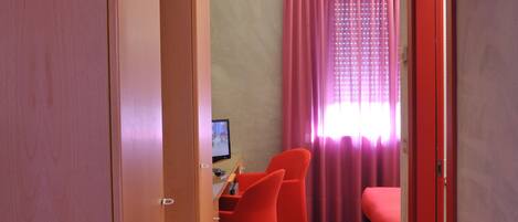 A hotel room with a red carpet, a desk with a computer, a bed, and a window with pink curtains.