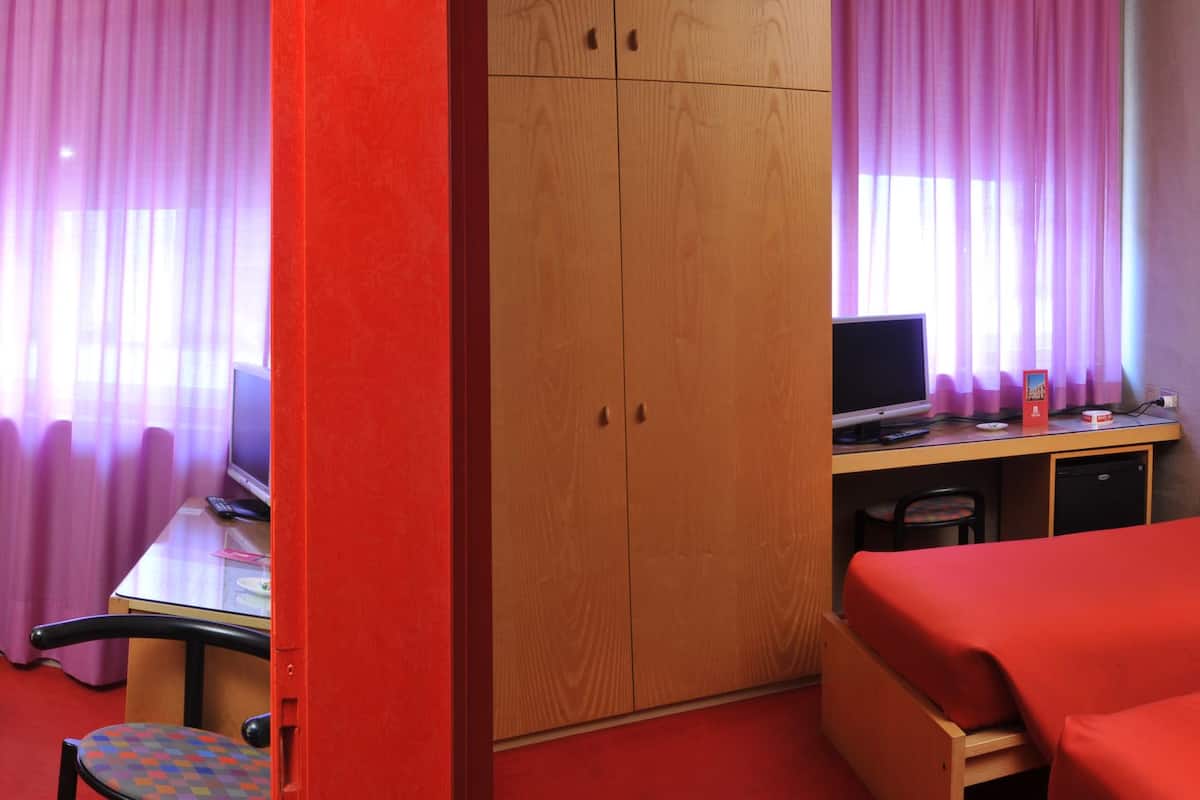 Quarto de hotel com duas camas de solteiro, uma escrivaninha com computador, uma mesinha e um guarda-roupa.