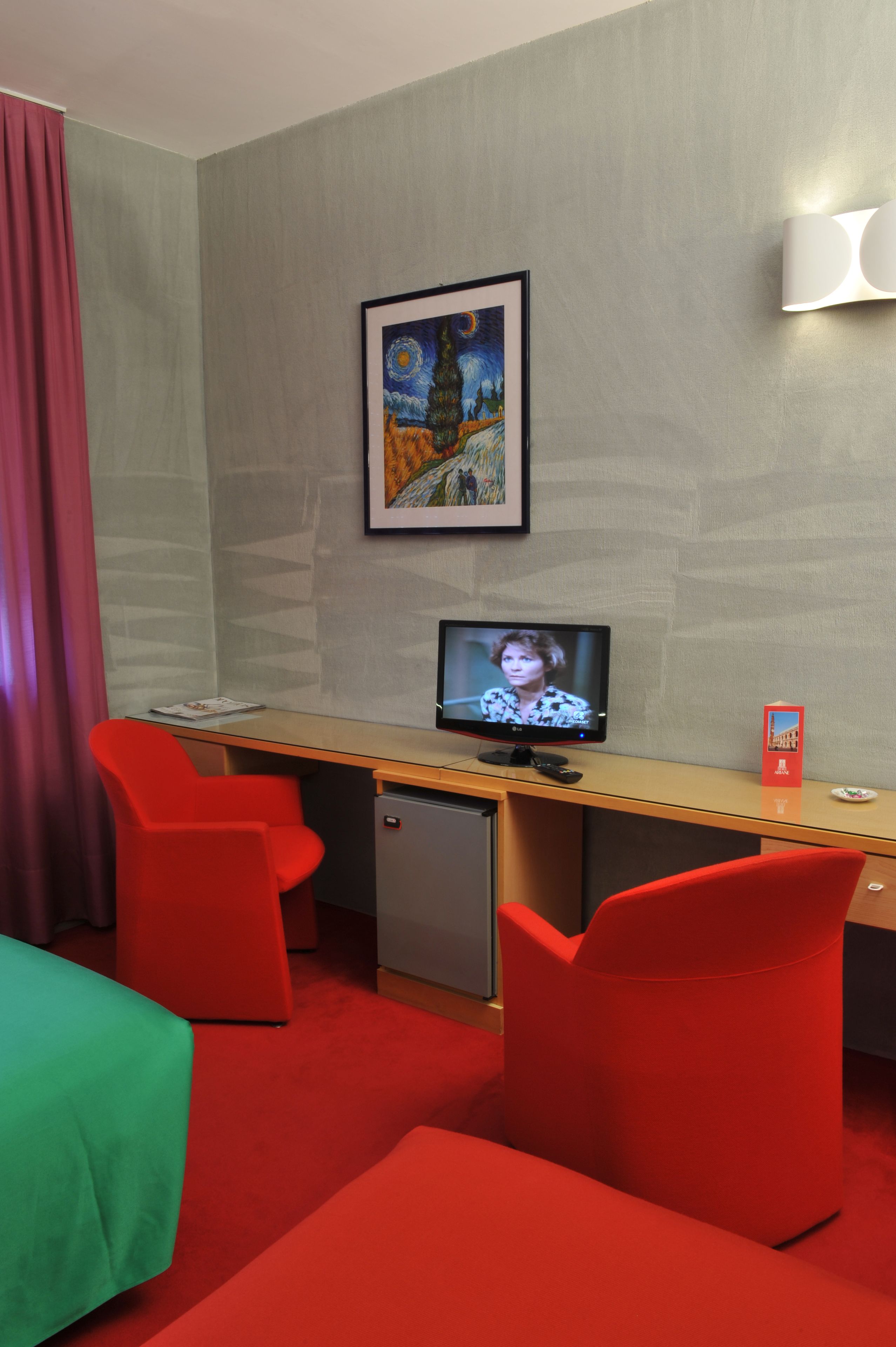 Ein Hotelzimmer mit Schreibtisch, Fernseher, Kühlschrank und roten Stühlen.