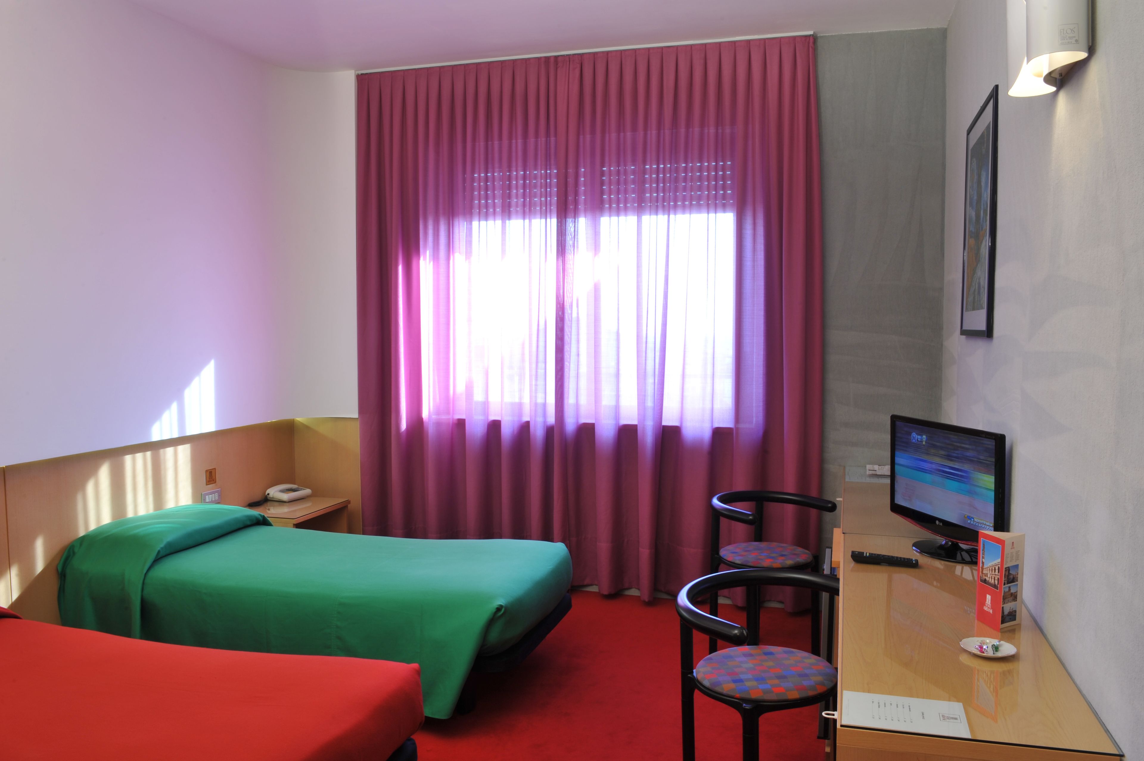 Una camera d'albergo con due letti, una scrivania con un computer, una sedia, una finestra con tende e una lampada fissata al muro.