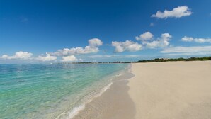 Une plage à l’eau turquoise et limpide, une vaste étendue de sable et, au loin, une côte bordée de bâtiments.