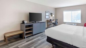 A modern hotel room with a large bed, a desk with a chair, a television, and a bench.