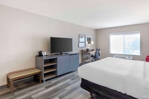 A modern hotel room with a large bed, a desk with a chair, a television, and a bench.