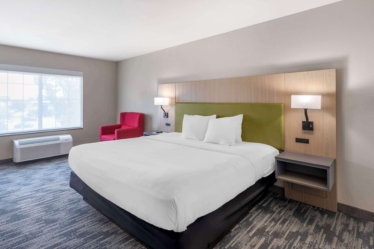 A hotel room with a large bed, two bedside lamps, a red chair, and a view of the outdoors through a large window.