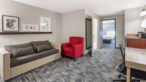 A hotel room with a sofa, a red armchair, a desk with a lamp, and framed artwork on the wall. - Country Inn & Suites by Radisson, St. Peters, MO (St. Charles)