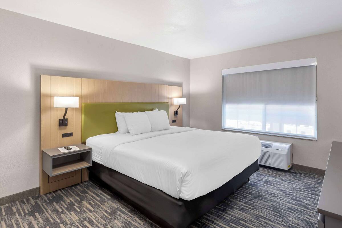A hotel room with a large bed, two bedside lamps, a nightstand, a built-in TV shelf, and a window with blinds.