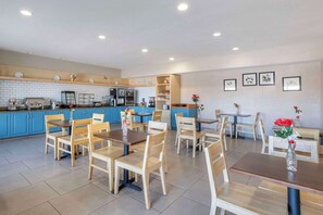 A spacious dining area with wooden tables and chairs, a kitchen in the background, and framed artwork on the walls.
