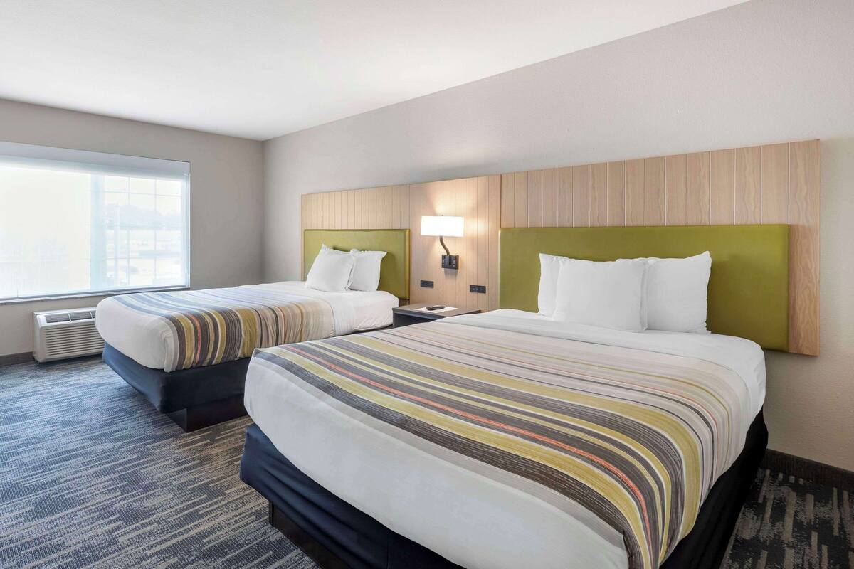 A hotel room with two beds, a large window, and a wall-mounted lamp.