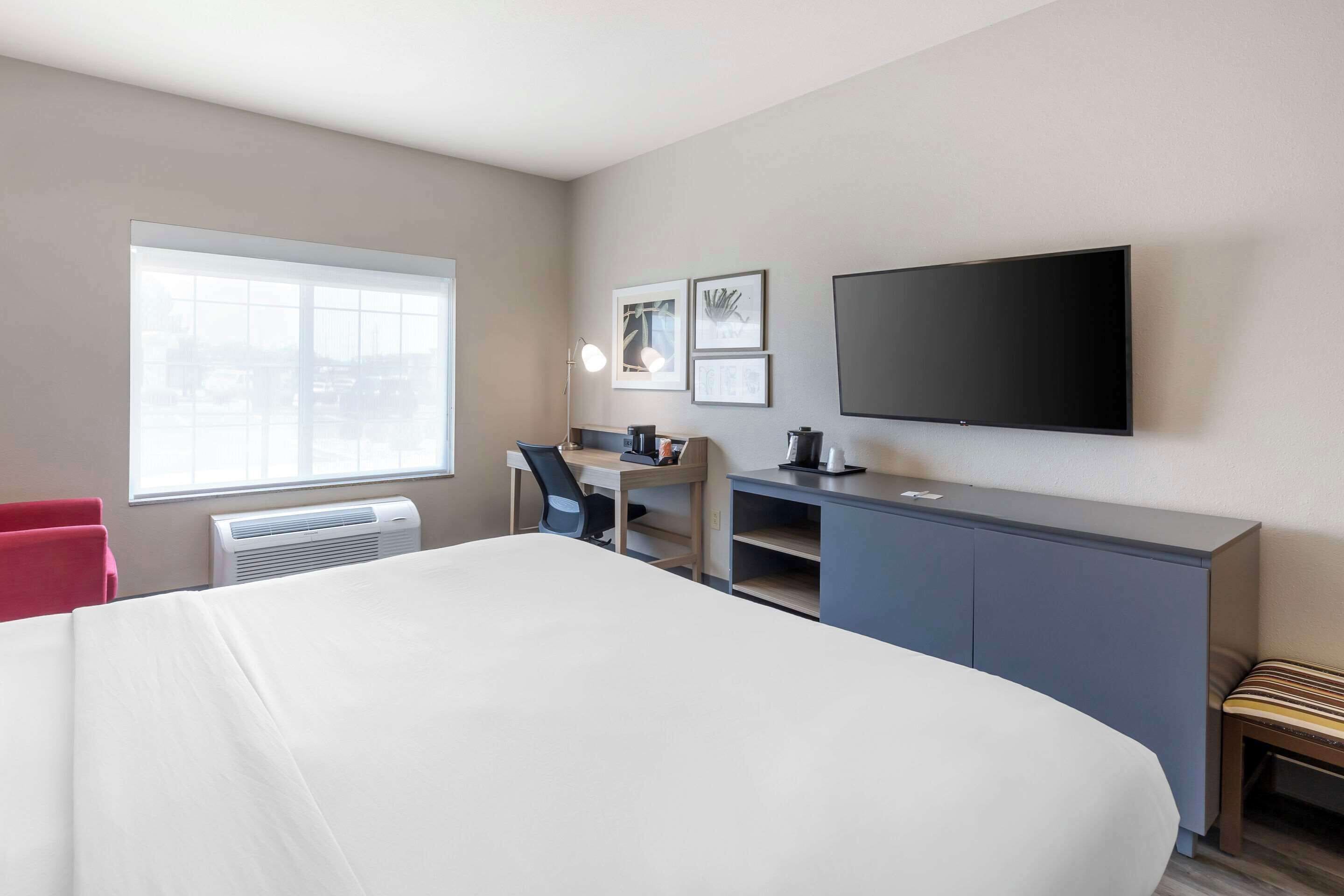 A hotel room with a large bed, a desk with a chair, a flat-screen TV mounted on the wall, and a window providing natural light.