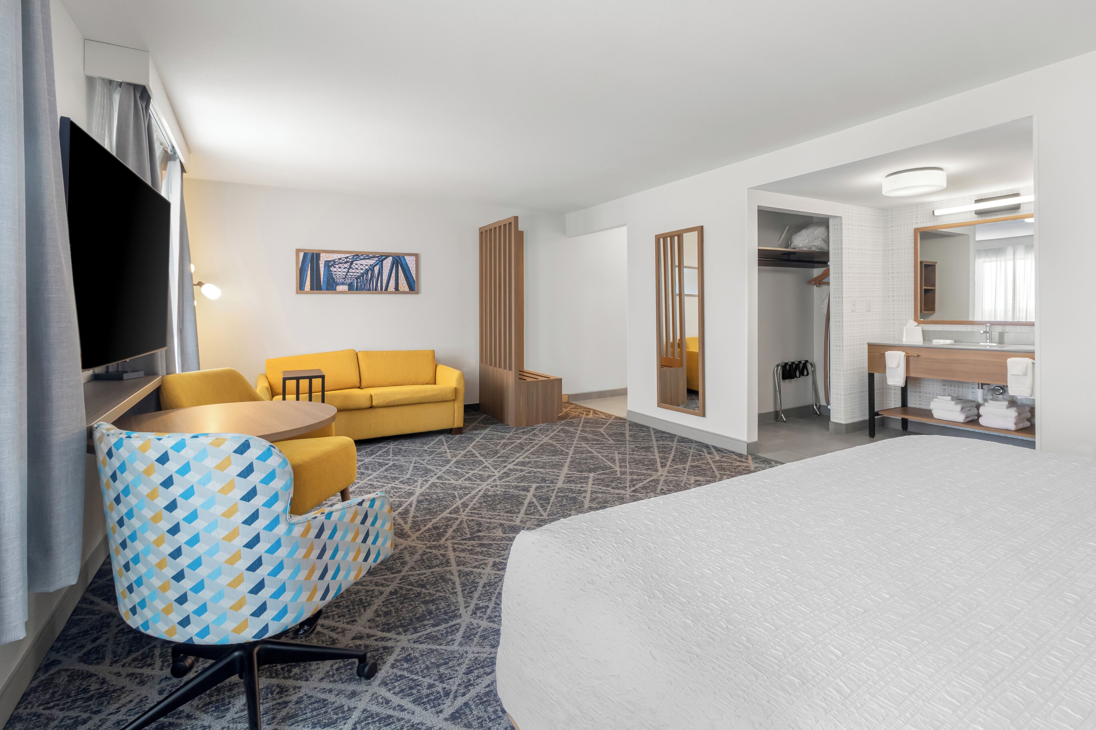A hotel room with a bed, a yellow sofa, a patterned armchair, a small round table, a TV, a painting on the wall, and a bathroom with a mirror and towel rack.