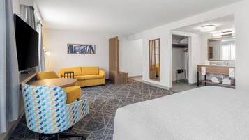 A hotel room with a bed, a yellow sofa, a patterned armchair, a small round table, a TV, a painting on the wall, and a bathroom with a mirror and towel rack.
