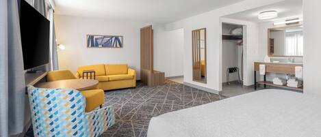 A hotel room with a bed, a yellow sofa, a patterned armchair, a small round table, a TV, a painting on the wall, and a bathroom with a mirror and towel rack.