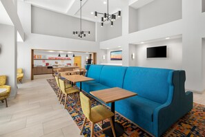 A modern hotel lobby with a blue sofa, wooden tables, yellow chairs, a kitchen area, and a flat-screen TV. - Hampton Inn & Suites New Castle (New Castle)