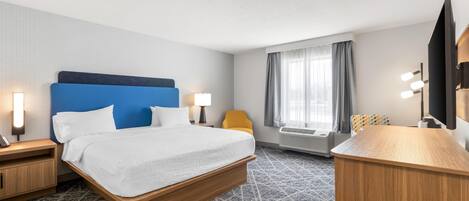 A hotel room with a bed, bedside tables, a desk, a chair, a window with curtains, an air conditioning unit, and a television.