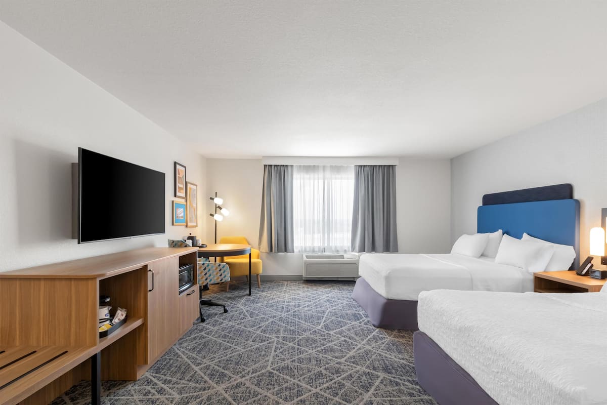 A hotel room with a flat-screen TV, two beds, a desk with a chair, a small table, and a window with curtains.