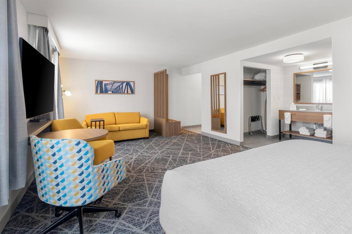 A hotel room with a bed, a sofa, a chair, a TV, a bathroom with a mirror, and a painting on the wall.