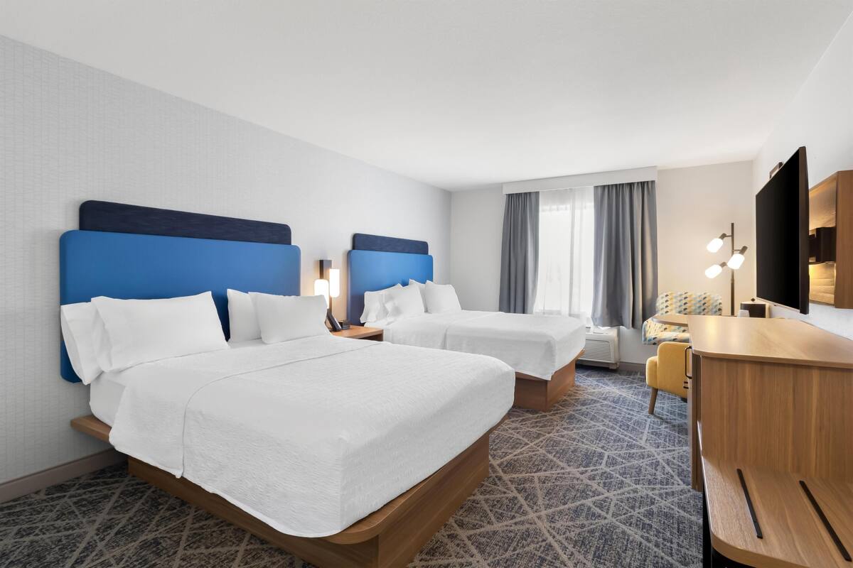 A hotel room with two beds, a desk, a chair, a TV, and a window with curtains.