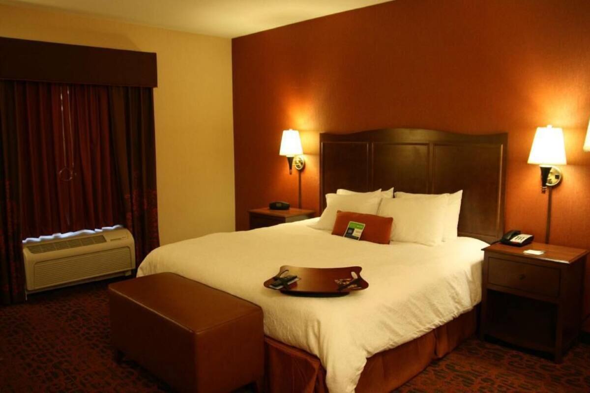 A hotel room with a large bed, two bedside lamps, a nightstand with a phone, a tray on the bed, a window with curtains, and an air conditioning unit.