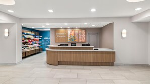 A hotel reception area with a wooden reception desk, a blue wall with a 'Welcome to Hampton' sign, and a snack display. - Hampton Inn & Suites New Castle (New Castle)