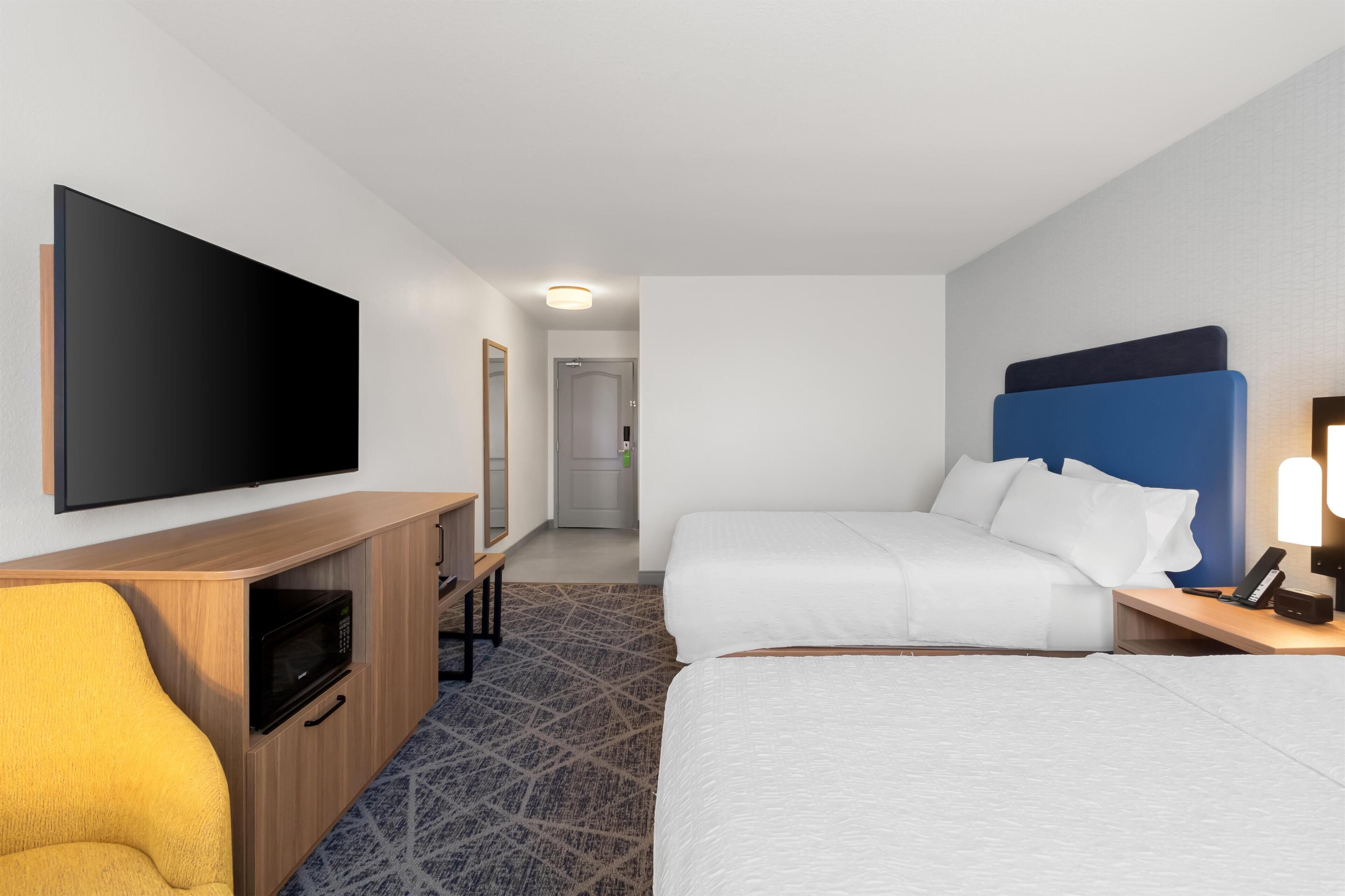 Quarto de hotel com televisão de tela plana, escrivaninha de madeira com micro-ondas, duas camas, mesa de cabeceira com abajur e carpete estampado.