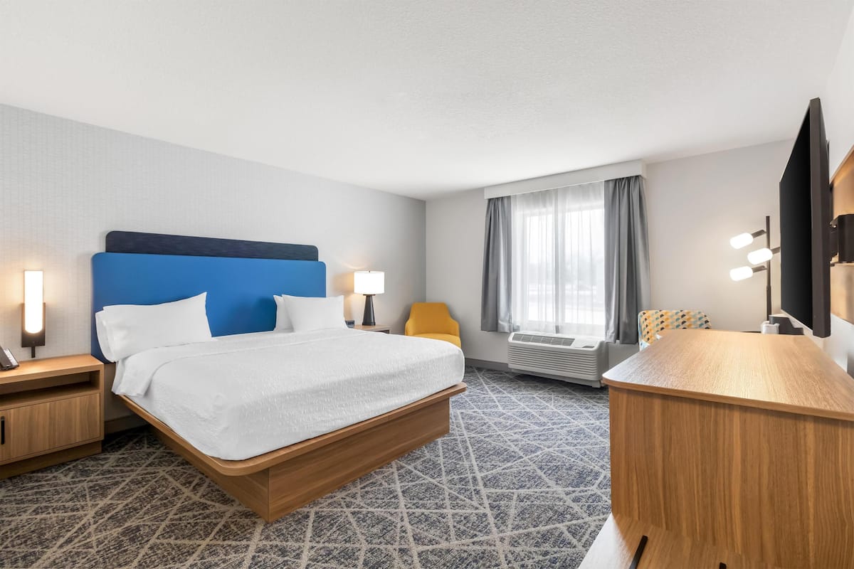A hotel room with a bed, bedside tables, a desk, a chair, an air conditioning unit, and a television.