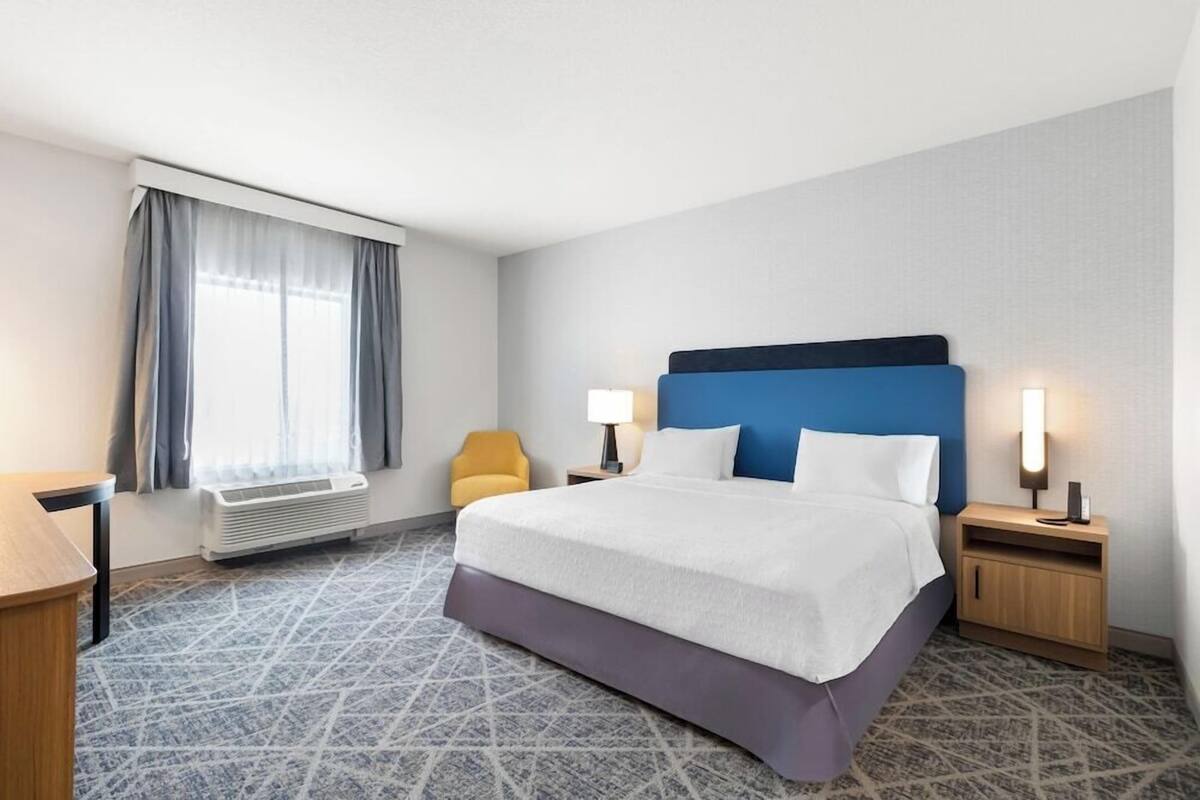 A hotel room with a bed, bedside tables, a chair, a window with curtains, and a heater.