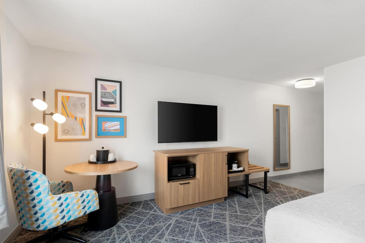 A hotel room with a bed, a TV, a microwave, a small table, a chair, and artwork on the wall.
