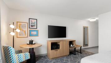 A hotel room with a bed, a TV, a microwave, a small table, a chair, and artwork on the wall.