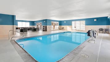A large indoor swimming pool with a blue interior, surrounded by a seating area with tables and chairs, and a view of the outdoors through large windows.
