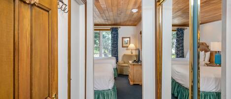 Een hotelkamer met twee bedden, een houten deur met nummerplaatje, een spiegel en een raam met gordijnen.