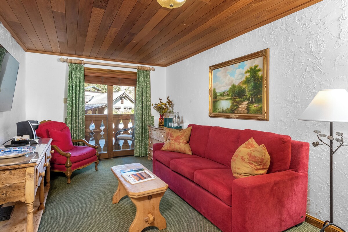 Una habitación con techo de madera, un sofá rojo, un escritorio de madera, una silla, un cuadro en la pared y una lámpara.
