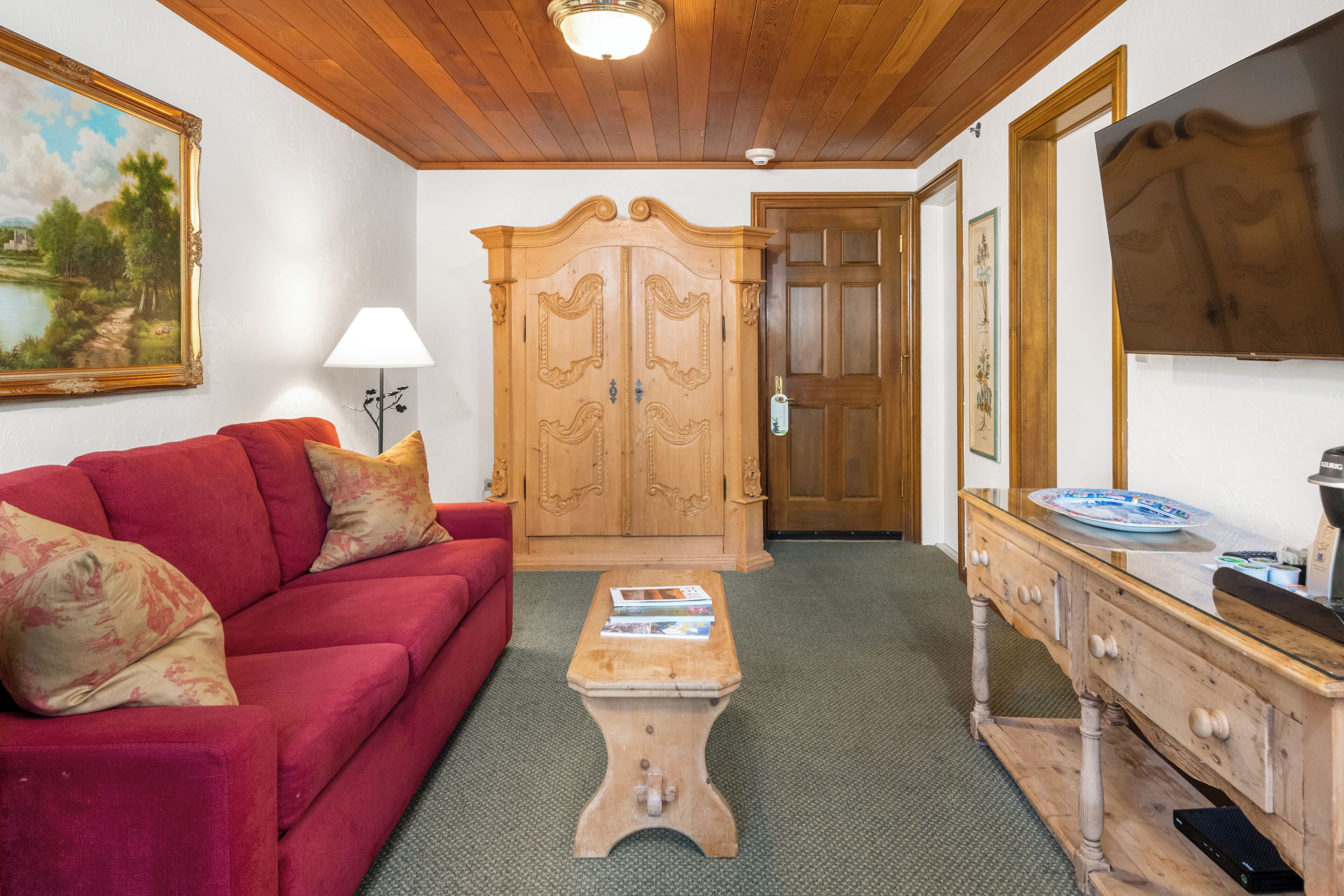 A room with a red sofa, a wooden cabinet, a small table with books, and a TV on the wall.