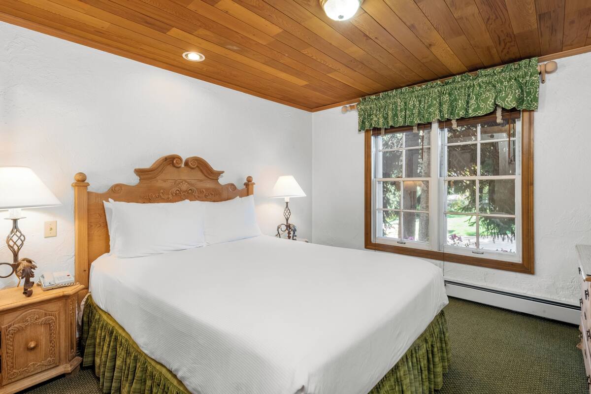 A neatly made bed with a wooden headboard, two bedside lamps, a window with a view of greenery, and a wooden ceiling.