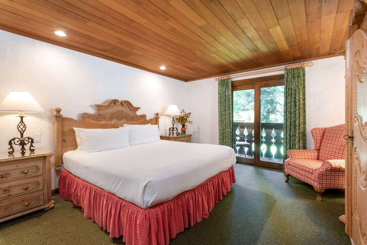 A bedroom with a wooden ceiling, a bed with a red checkered skirt, a wooden nightstand, a chair, and a view through a glass door.
