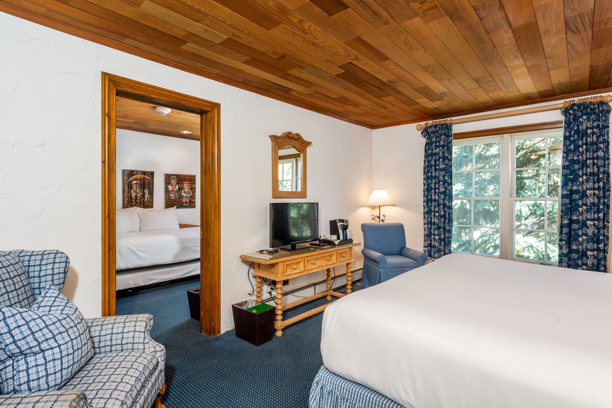 Habitación de hotel con techo de madera, una cama, un escritorio con televisor, una silla y una ventana con cortinas.