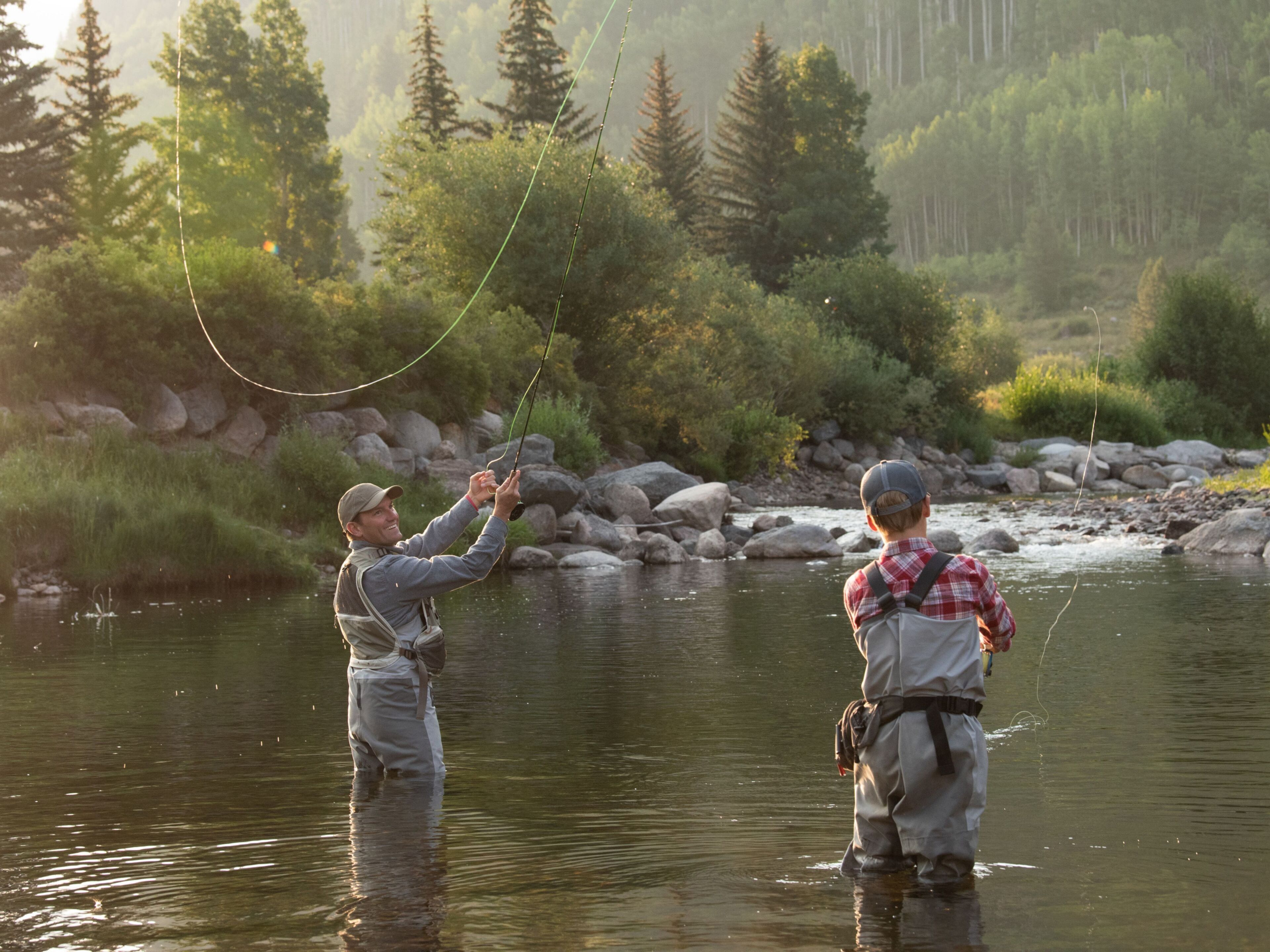 Two people fishing in a river surrounded by rocks and trees.