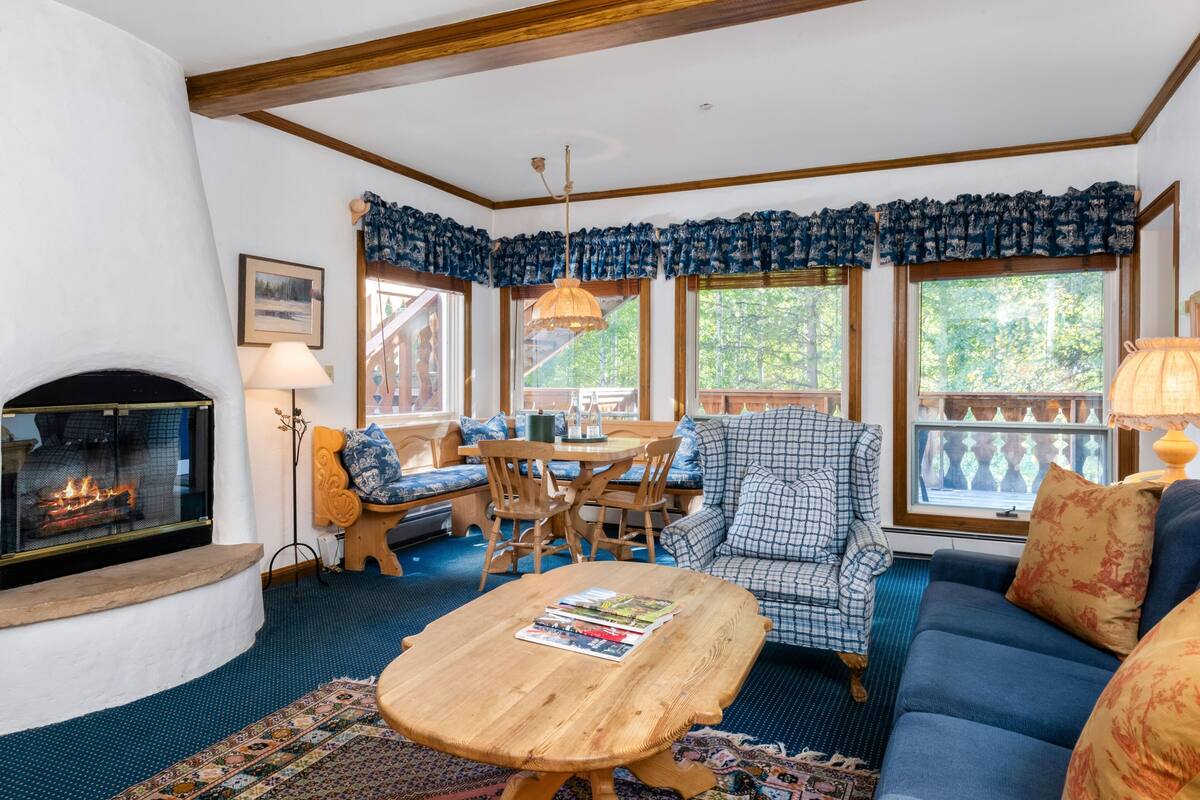 A cozy living room with a fireplace, wooden furniture, and large windows.